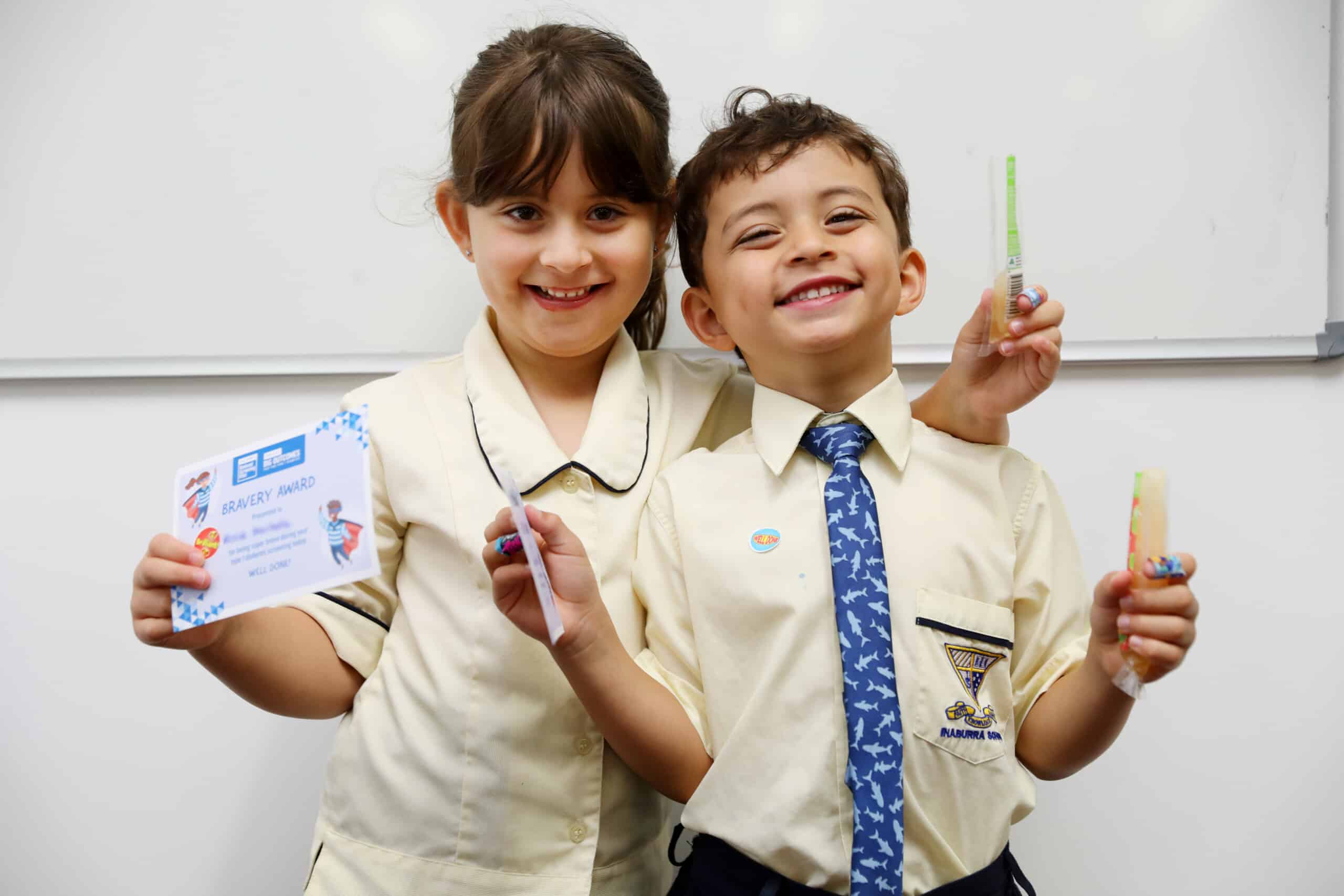 Two children smiling happily at the camera after participating in the T1D National Screening Pilot. They are wearing school uniforms and holding ice blocks and certificates for their participation.