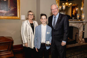 12 year-old JDRF advocate Nikolas (centre) in white shirt and blue blazer holding award. WA Governor stands on his right in a black suit, with his wife on the left in a white and black striped blazer. 