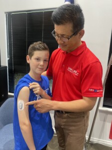 Nikolas Mirco stands on the left in a JDRF blue t-shirt. He holds his sleeve up to show his CGM. Sam Lim MP stands on the right in a red polo t-shirt, pointing at Nik's arm. 