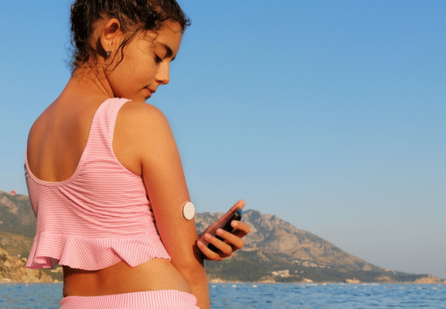 young women checking BGLs on a sensor on the back of her arm, wearing swimmers, water landscape in the background